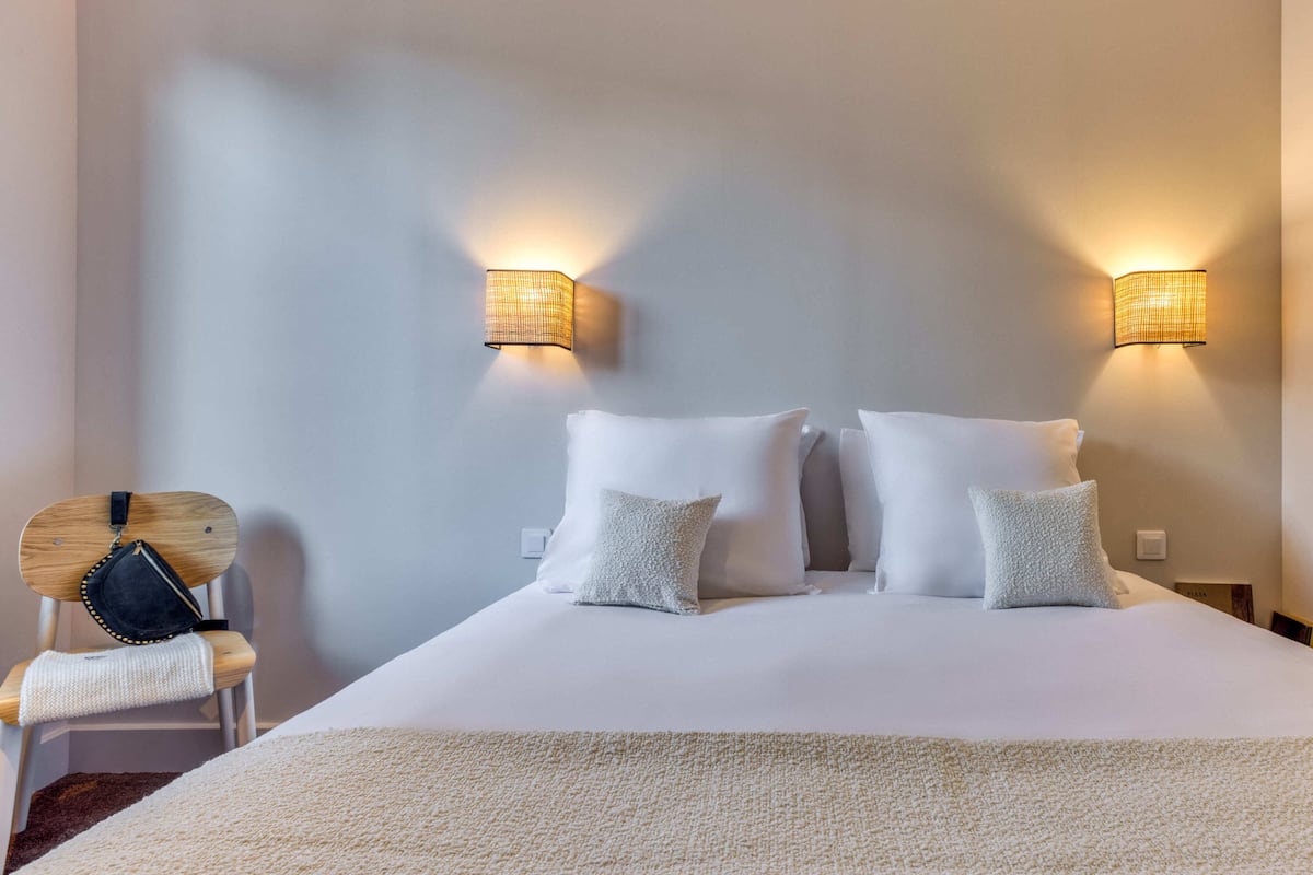 Un lit bien fait, avec du linge de lit blanc et des oreillers, deux lampes fixées au mur et une chaise en bois sur laquelle est posé un sac noir.