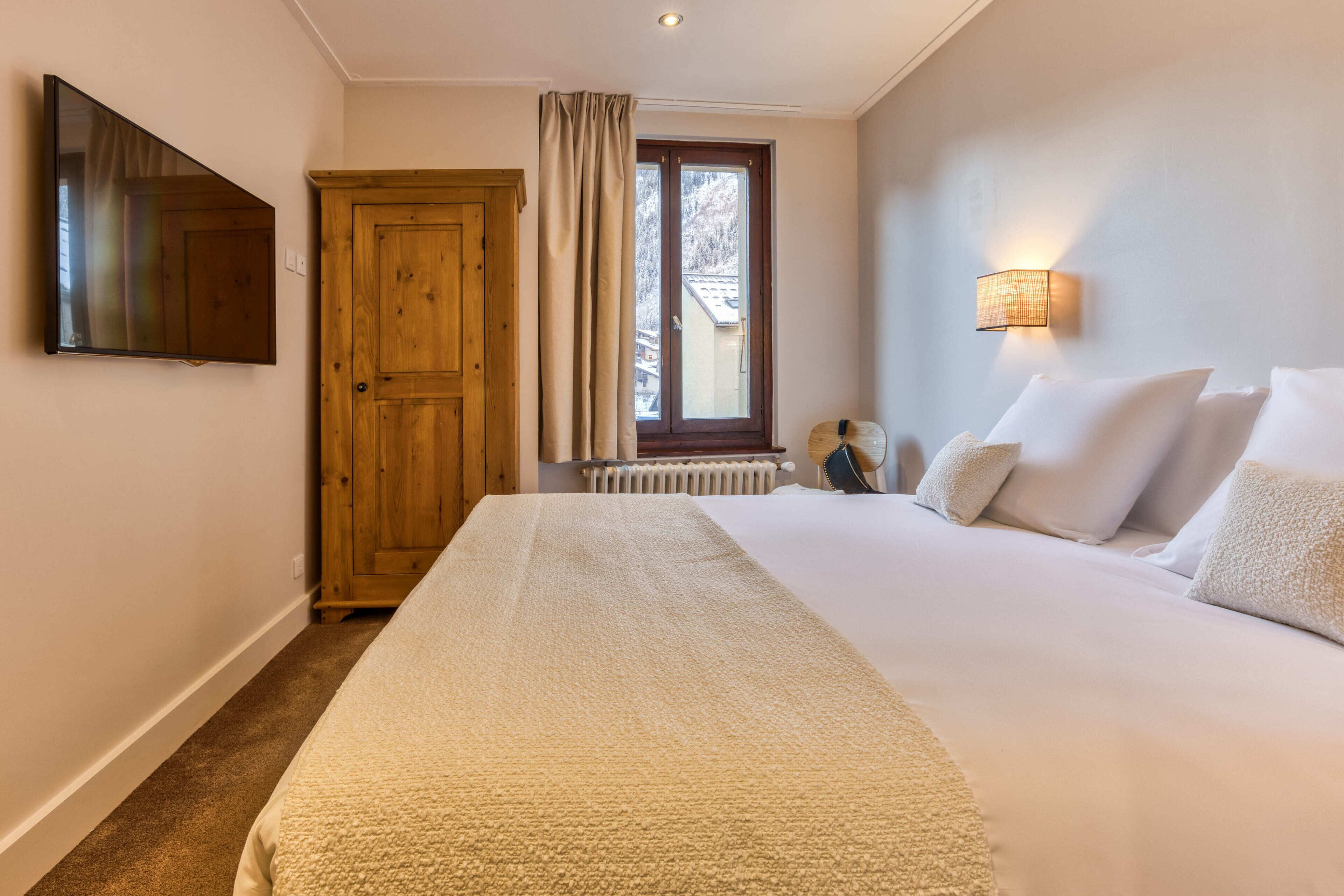 Une chambre d’hôtel avec un grand lit, une armoire en bois, un téléviseur fixé au mur et une fenêtre avec des rideaux.