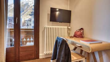A hotel room with a wooden desk, a television mounted on the wall, a radiator, and a view of snow-capped mountains through a glass door.