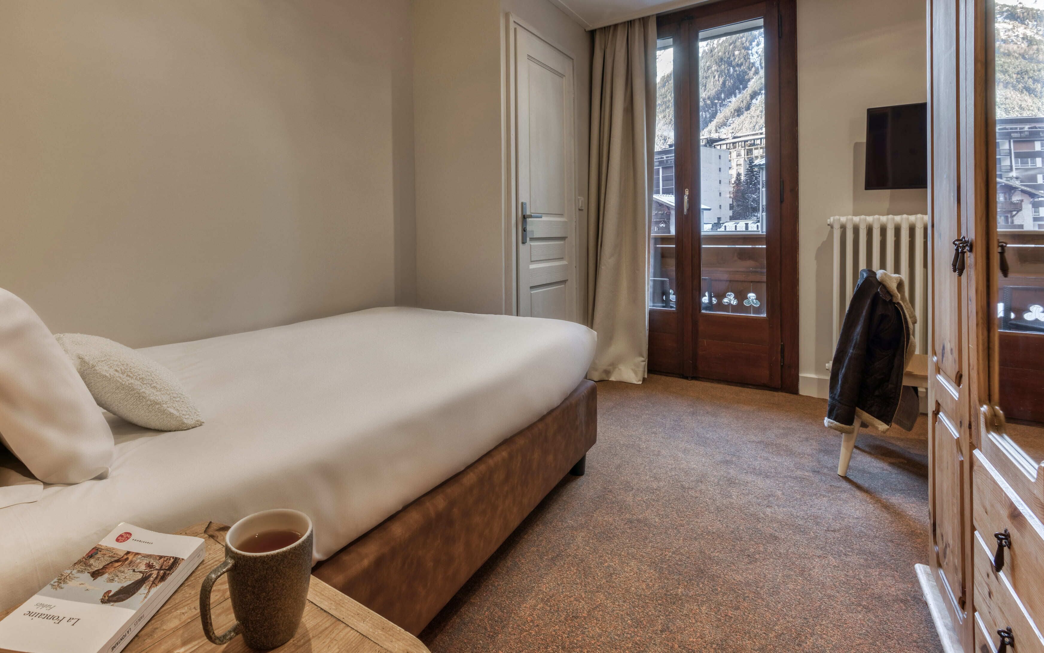 Une chambre à coucher comprenant un lit, une commode en bois, un radiateur et une fenêtre donnant sur un immeuble et des arbres.