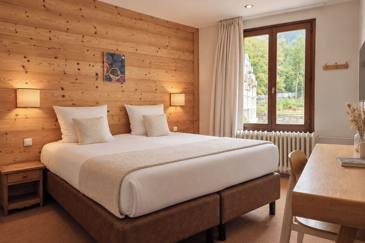 Een slaapkamer met een houten hoofdbord, een groot bed met witte lakens, bedlampen, een raam met uitzicht en een houten tafel met stoelen.