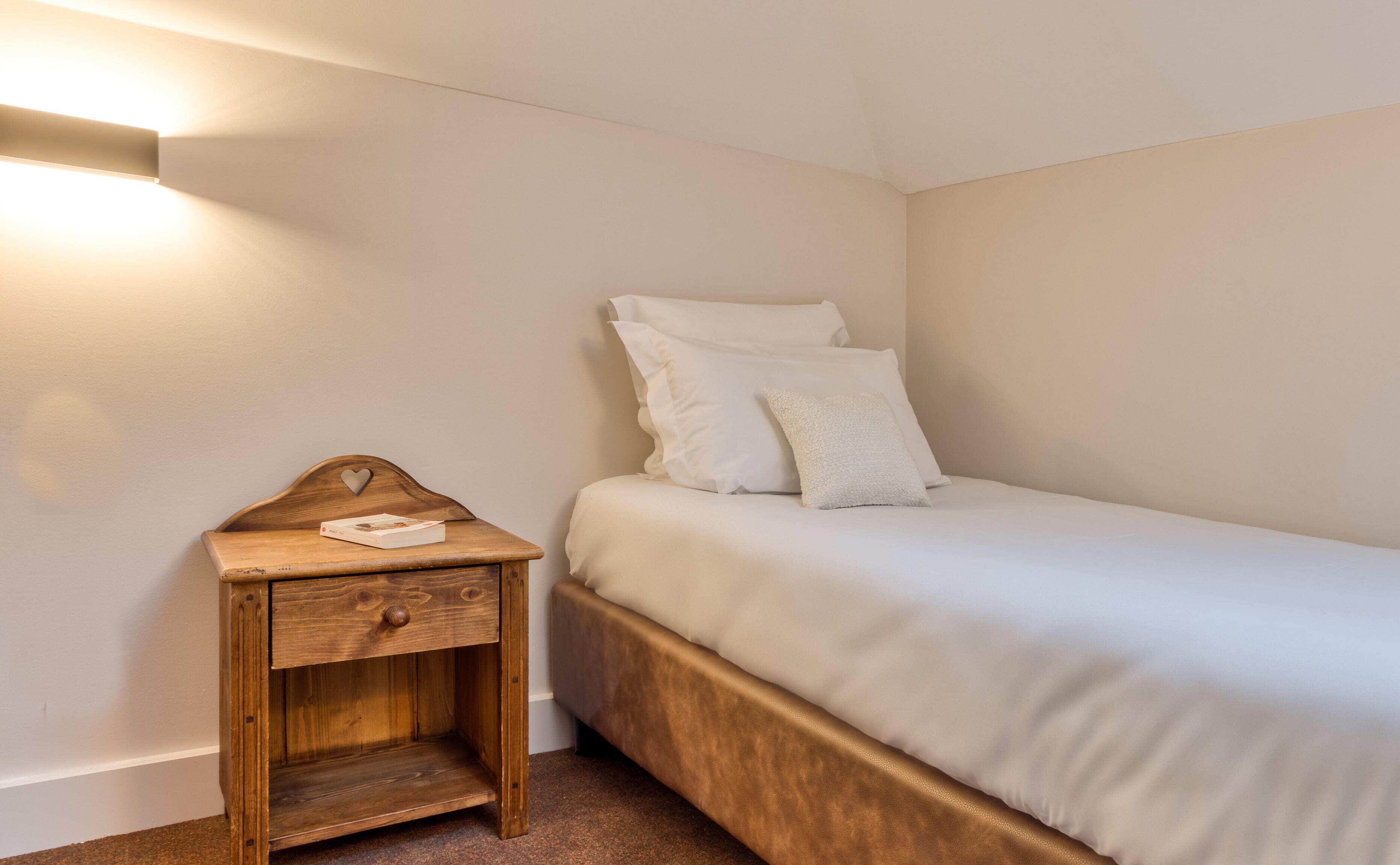A small, neatly made bed with white bedding and pillows, a wooden nightstand with a drawer, and a wall-mounted light fixture.