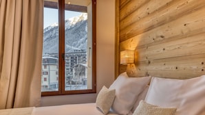 Habitación de hotel con cabecera de madera, cama con ropa blanca y vistas a montañas nevadas a través de una ventana.