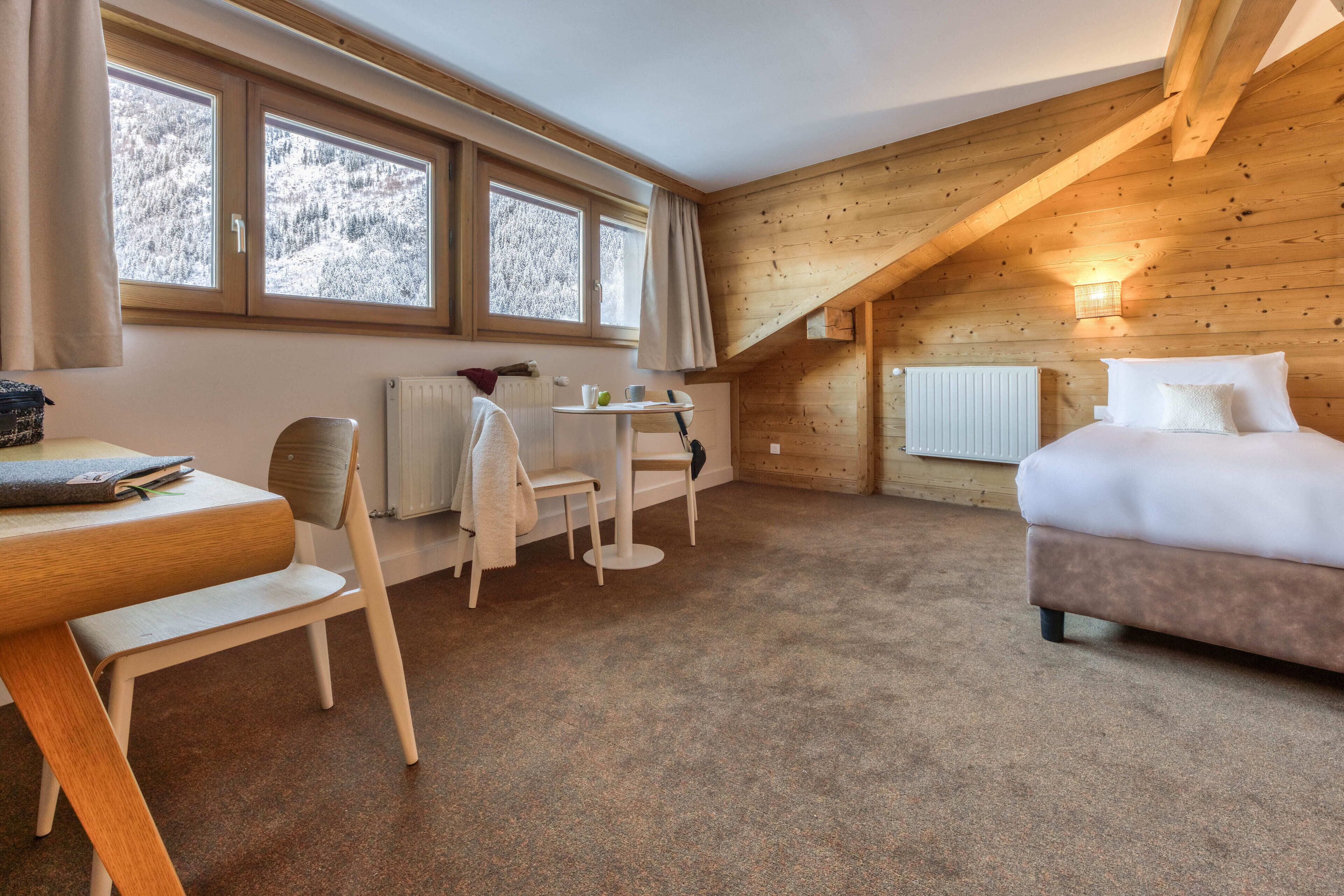 A cozy room with a bed, a desk, and a chair. There are windows with curtains, a radiator, and a lamp.