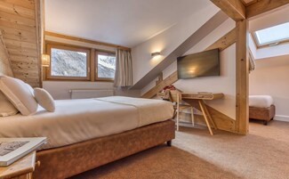 A bedroom with a bed, a desk, a chair, a TV, and a window.