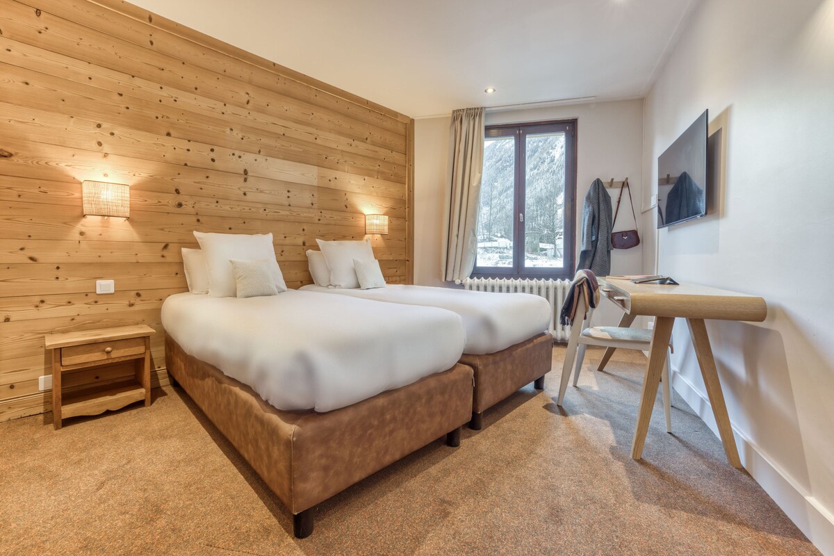 Une chambre avec des lambris en bois, un lit double, une table de chevet, une chaise, un bureau et une fenêtre donnant sur des arbres.