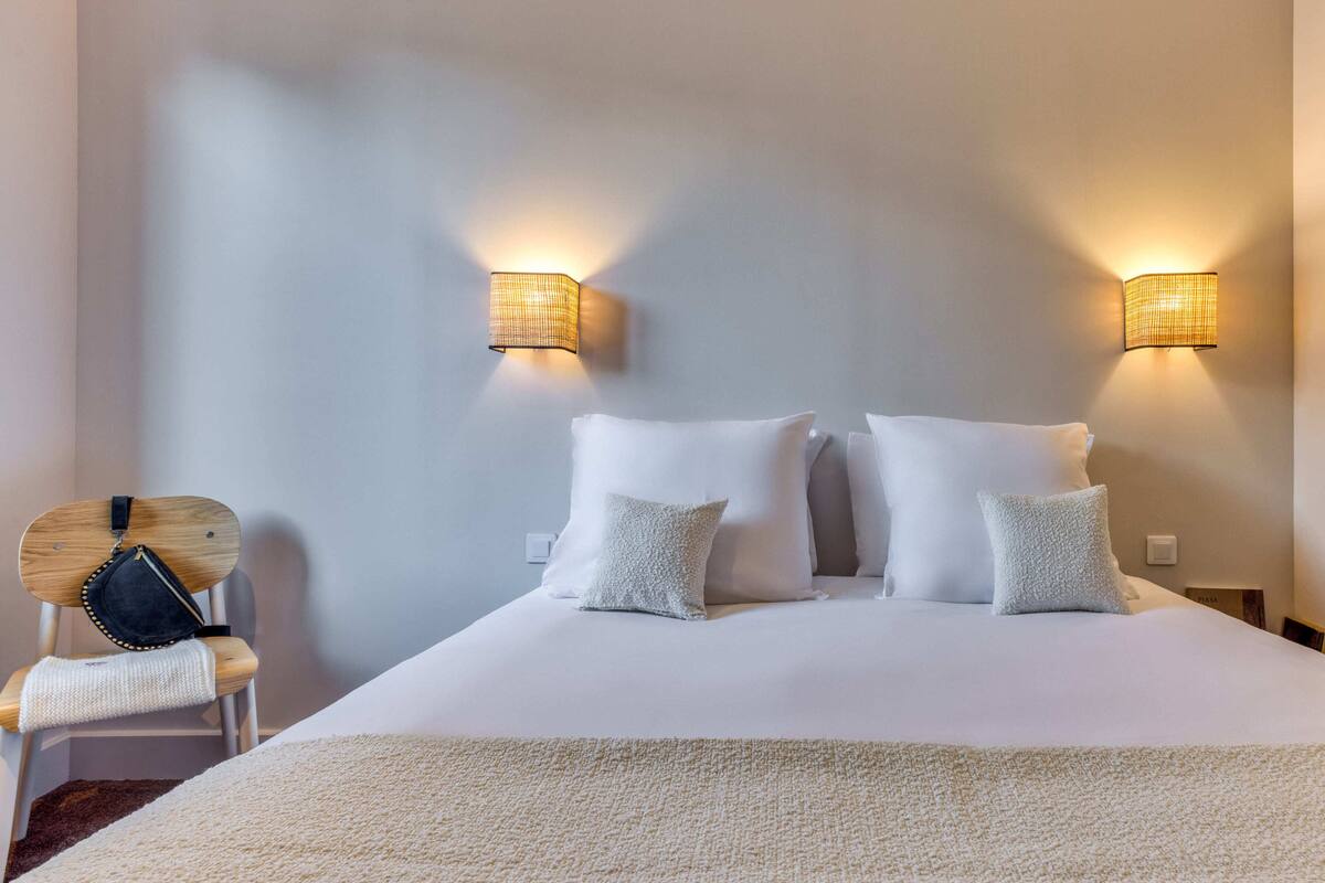 Un lit bien fait, avec du linge de lit blanc et des oreillers, deux lampes fixées au mur et une chaise en bois sur laquelle est posé un sac noir.