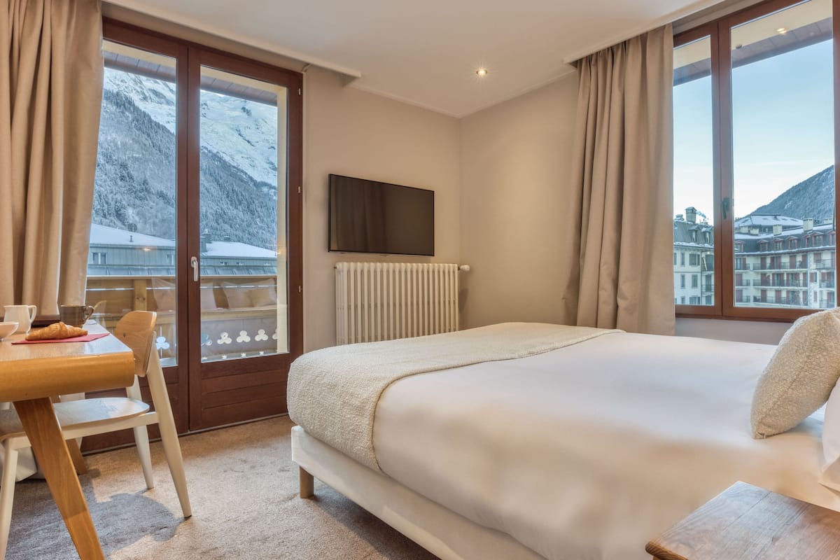 Une chambre d’hôtel avec un lit, un bureau et une chaise, un téléviseur fixé au mur et une vue sur des montagnes enneigées, visible à travers de grandes fenêtres.