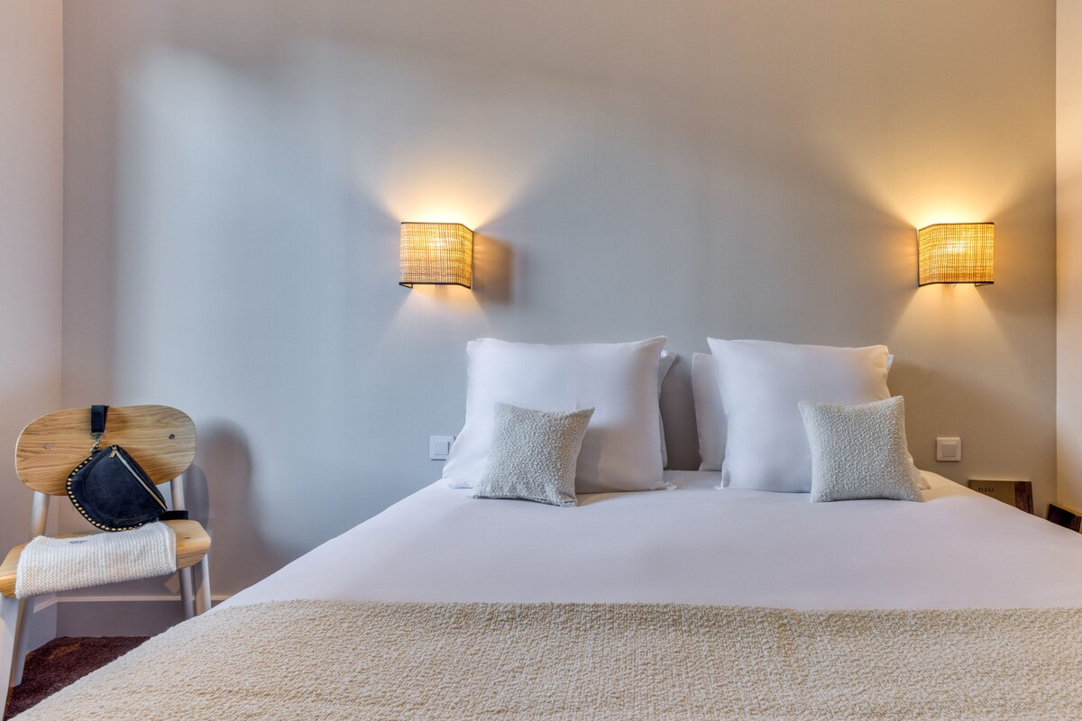 Un lit bien fait, avec du linge de lit blanc et des oreillers, deux lampes fixées au mur et une chaise en bois sur laquelle est posé un sac noir.