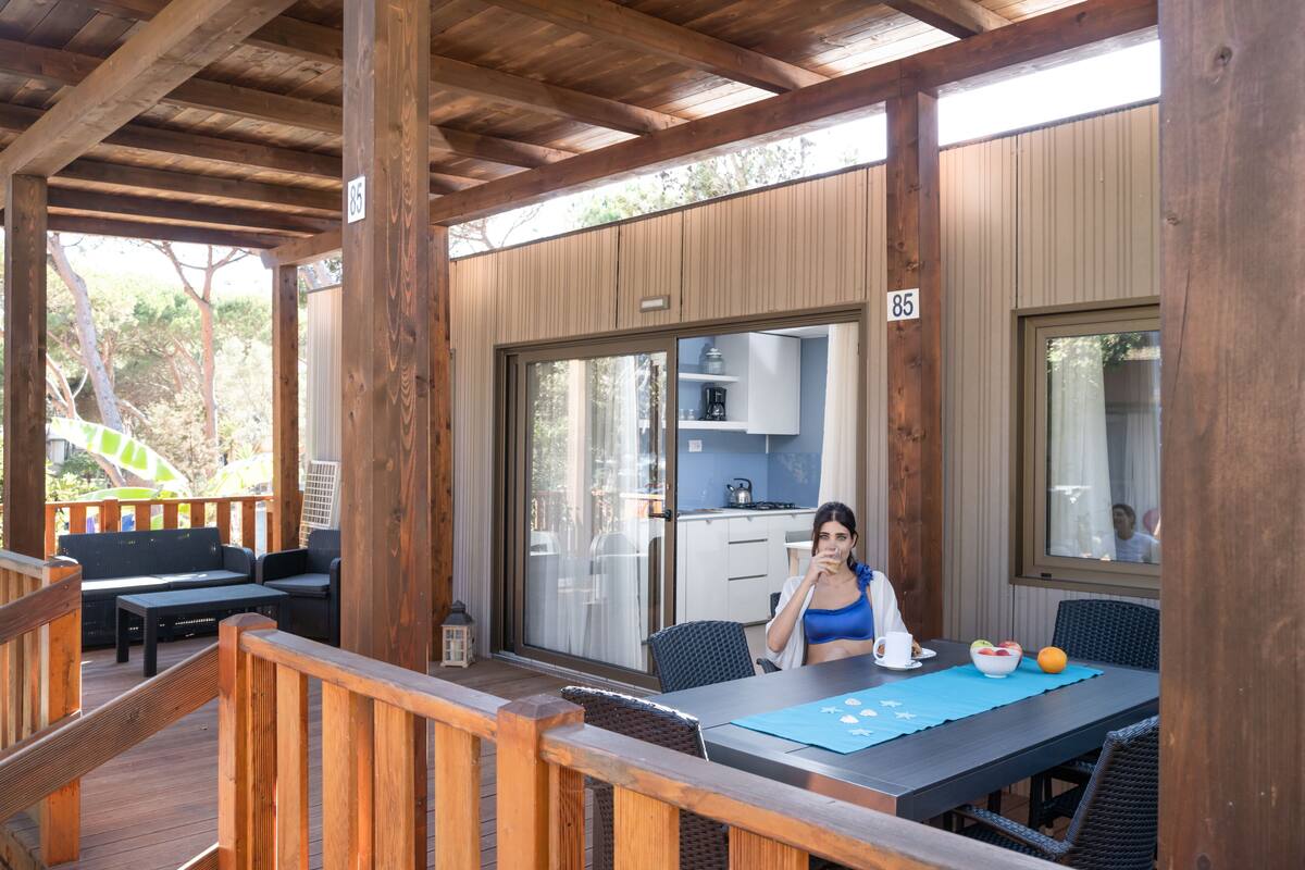 Un espacio comedor al aire libre con techo, un piso de madera, una mesa con sillas y vista a una cocina a través de una puerta de vidrio.