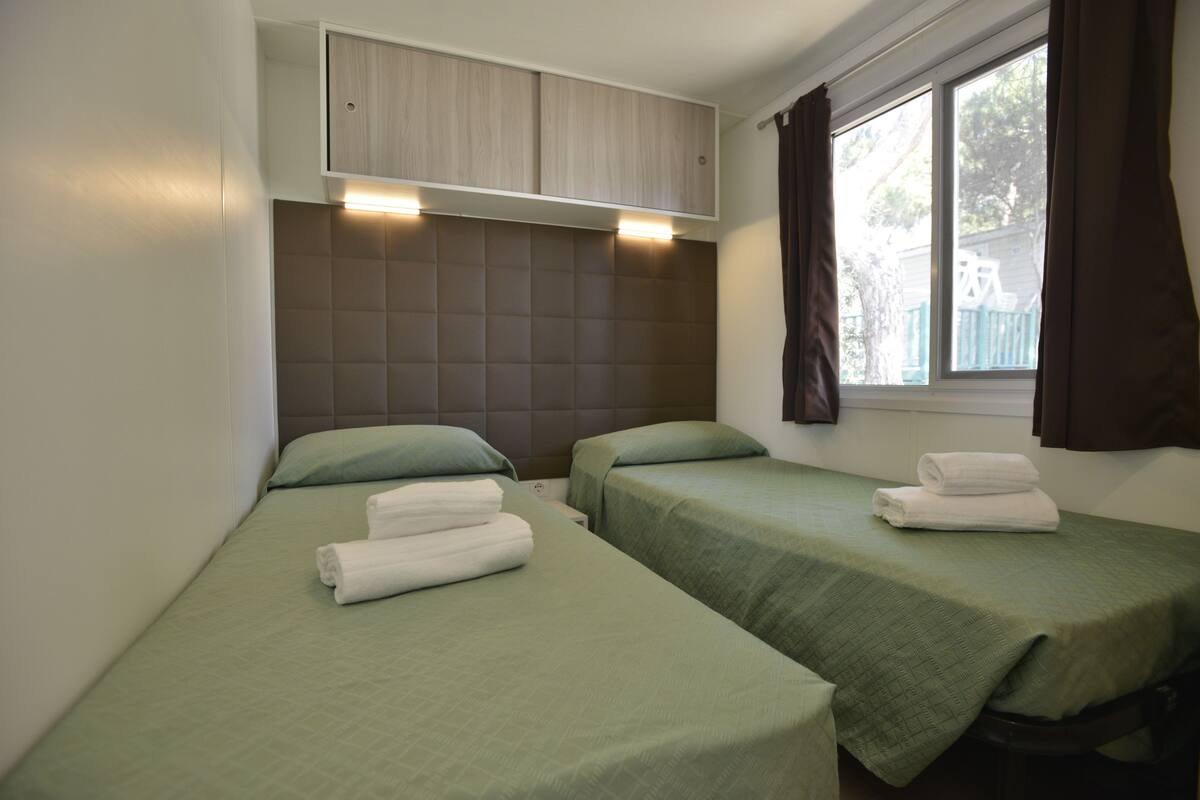 Una habitación con dos camas individuales, cada una con cubrecama verde y toallas blancas. Se ve una ventana con cortinas marrones, y un cabecero empotrado con una lámpara sobre las camas.