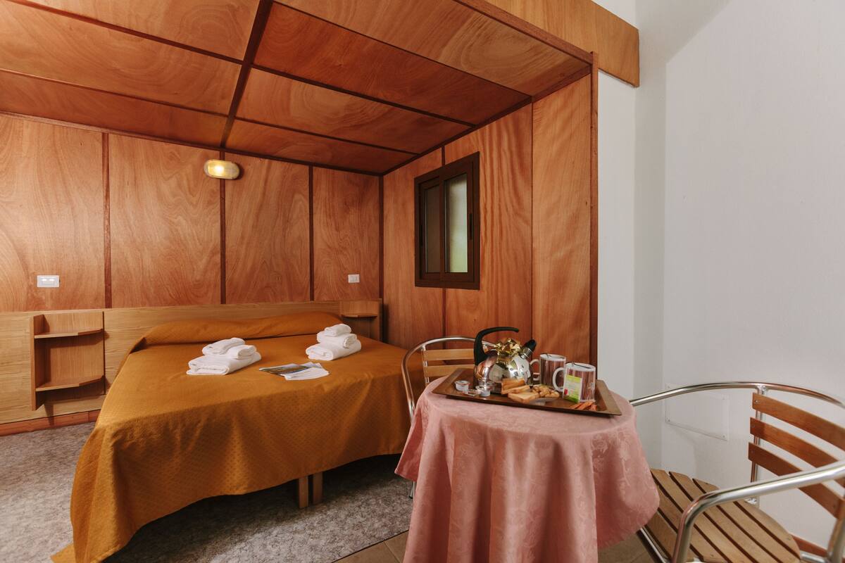 Una habitación con techo de madera, cama con colcha marrón, mesa con mantel rosa y una silla.