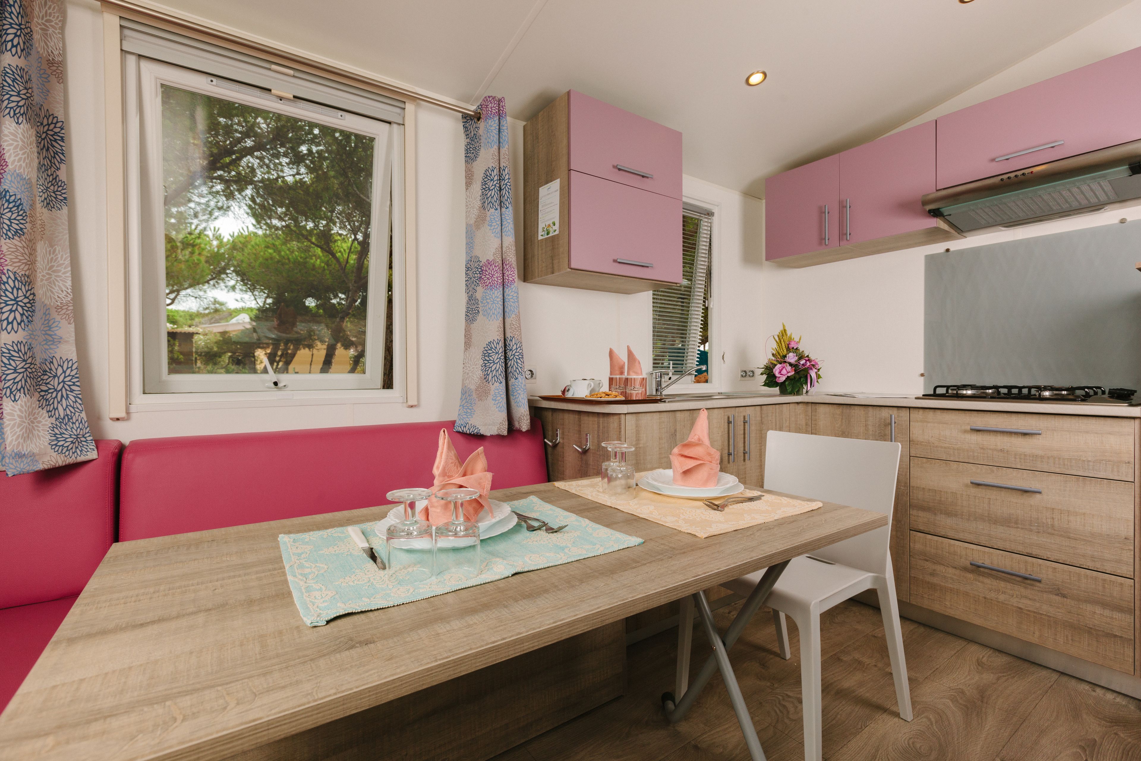 Un coin repas avec une table en bois, des chaises roses et blanches, et une fenêtre donnant sur les arbres.
