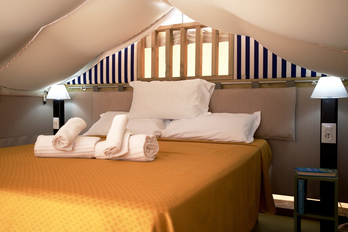 A hotel room with a bed, pillows, rolled towels, a nightstand with a lamp, and a small table with a book.
