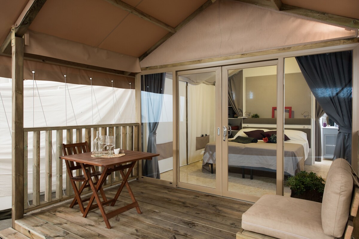 A wooden deck with a table and chairs, a sliding glass door leading to a bedroom with a bed and a small table.