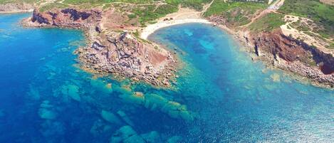 An aerial view of a coastal area with rocky terrain, clear blue waters, and a sandy beach.