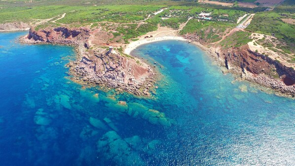 Vista aérea de una zona costera con terreno rocoso, aguas azul cristalina y una playa arenosa.