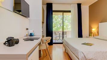 Ein Hotelzimmer mit einem Bett, einem Schreibtisch mit einer Kamera, einem Sessel, einem Tisch, einem Fenster mit Vorhängen und Blick auf Grünfläche.