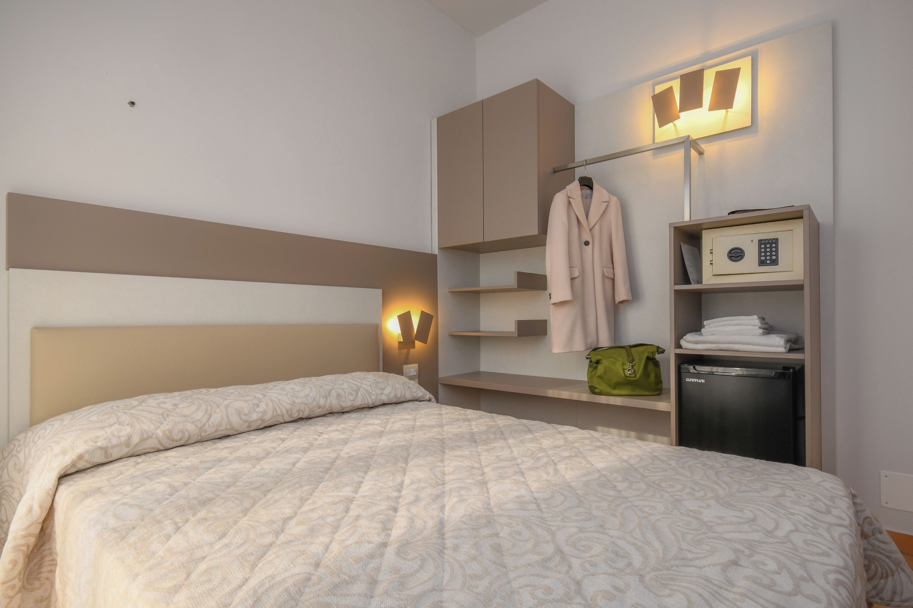 A neatly made bed with a patterned bedspread, a wardrobe with a hanging coat, a shelf with folded towels, and a small refrigerator.