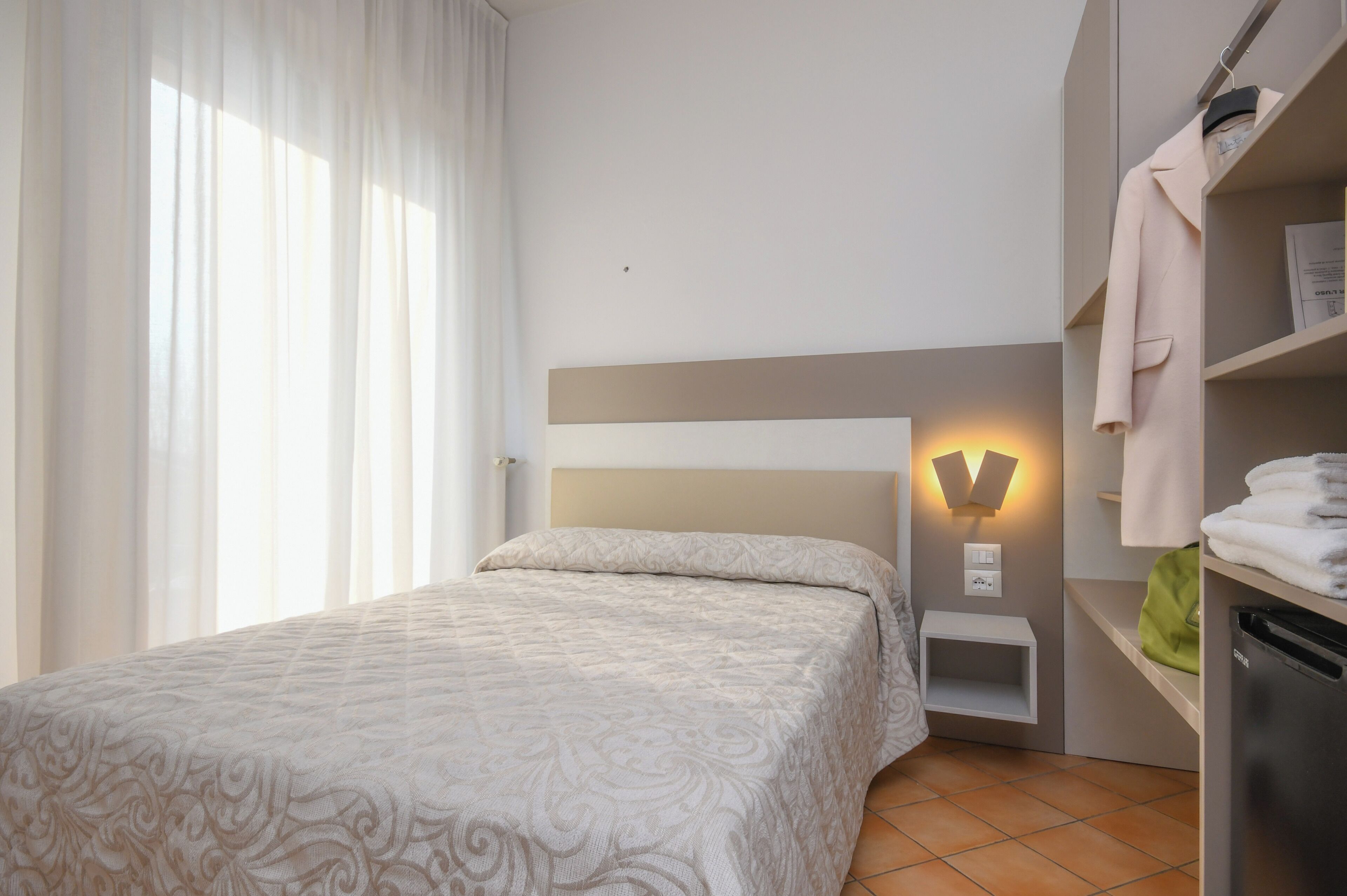 Un dormitorio con cama, mesita de noche, lámpara de pared, armario y ventana con cortinas.