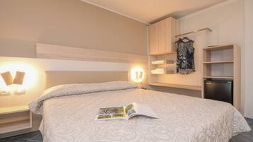 A neatly made bed with a patterned bedspread, a magazine on it, and a wall-mounted shelf with folded towels.