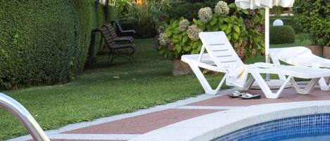 Una piscina con una escalera metálica, sillas de descanso blancas y una sombrilla.