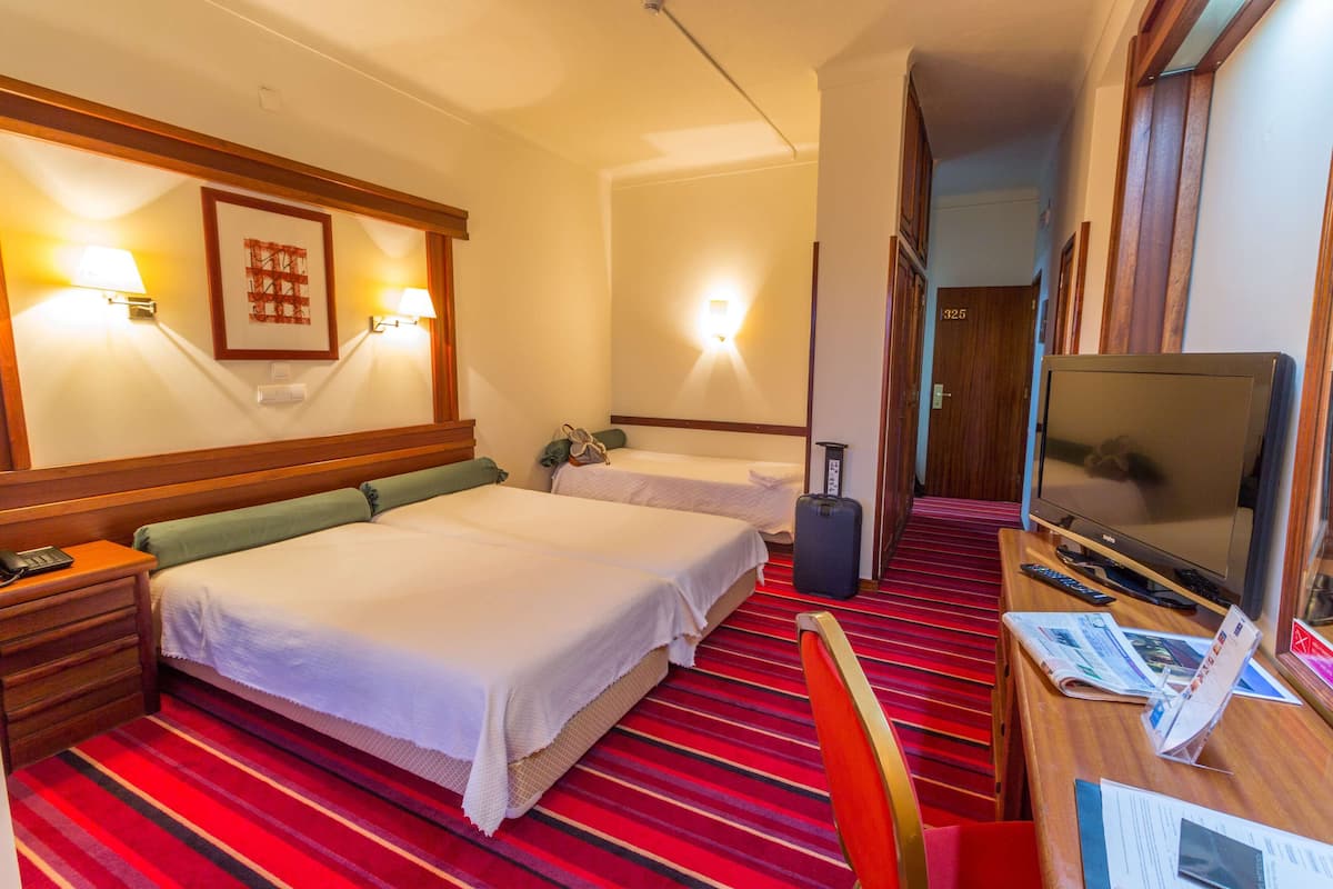 A hotel room with two beds, a desk with a TV, a suitcase, and a red striped carpet.