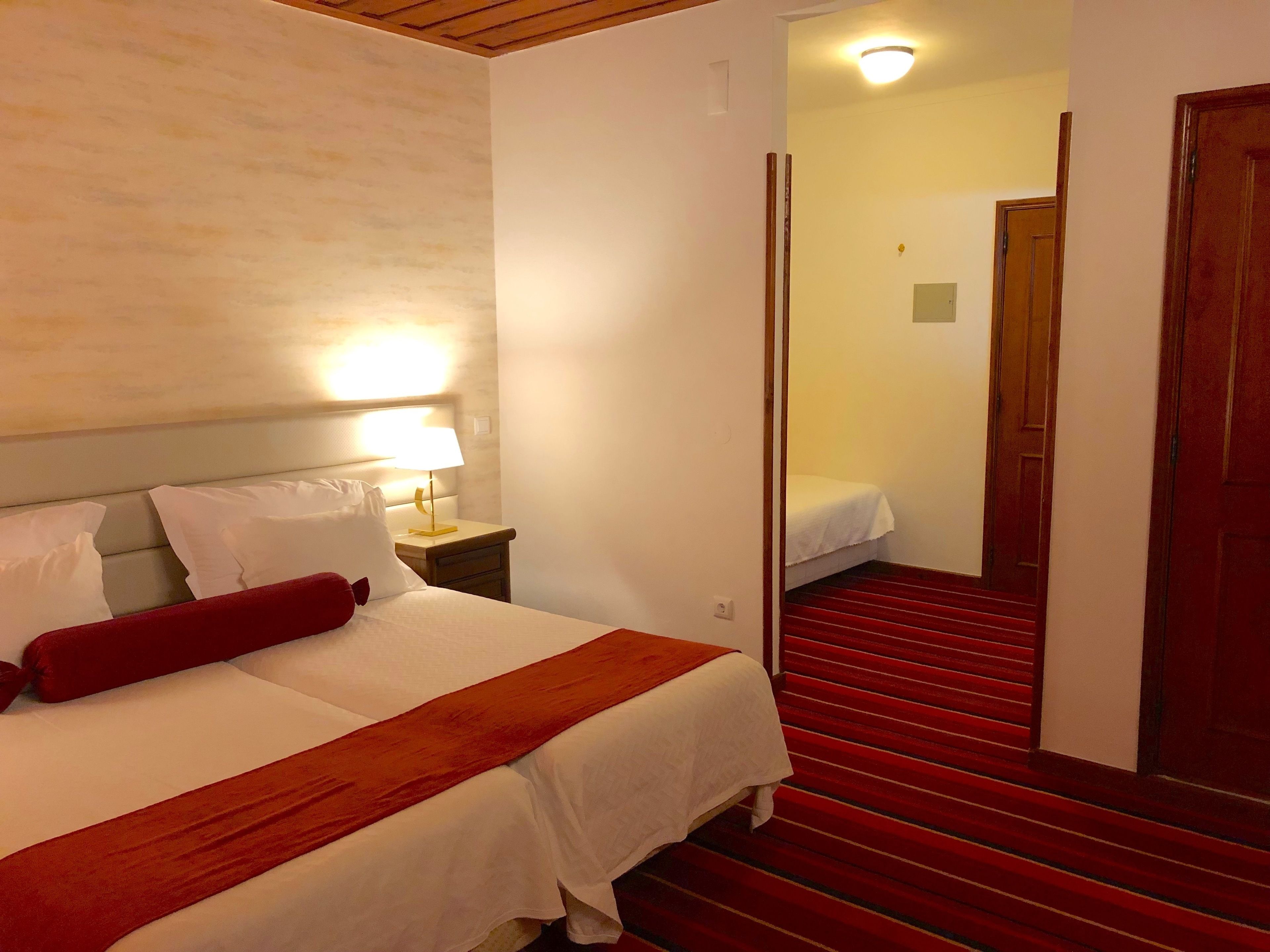 A hotel room with a bed, a nightstand, a lamp, a red carpeted staircase, and a door leading to another room.