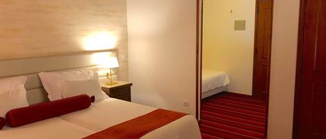 Quarto de hotel com cama, mesa de cabeceira, abajur, escada com carpete vermelho e porta que dá para outro aposento.