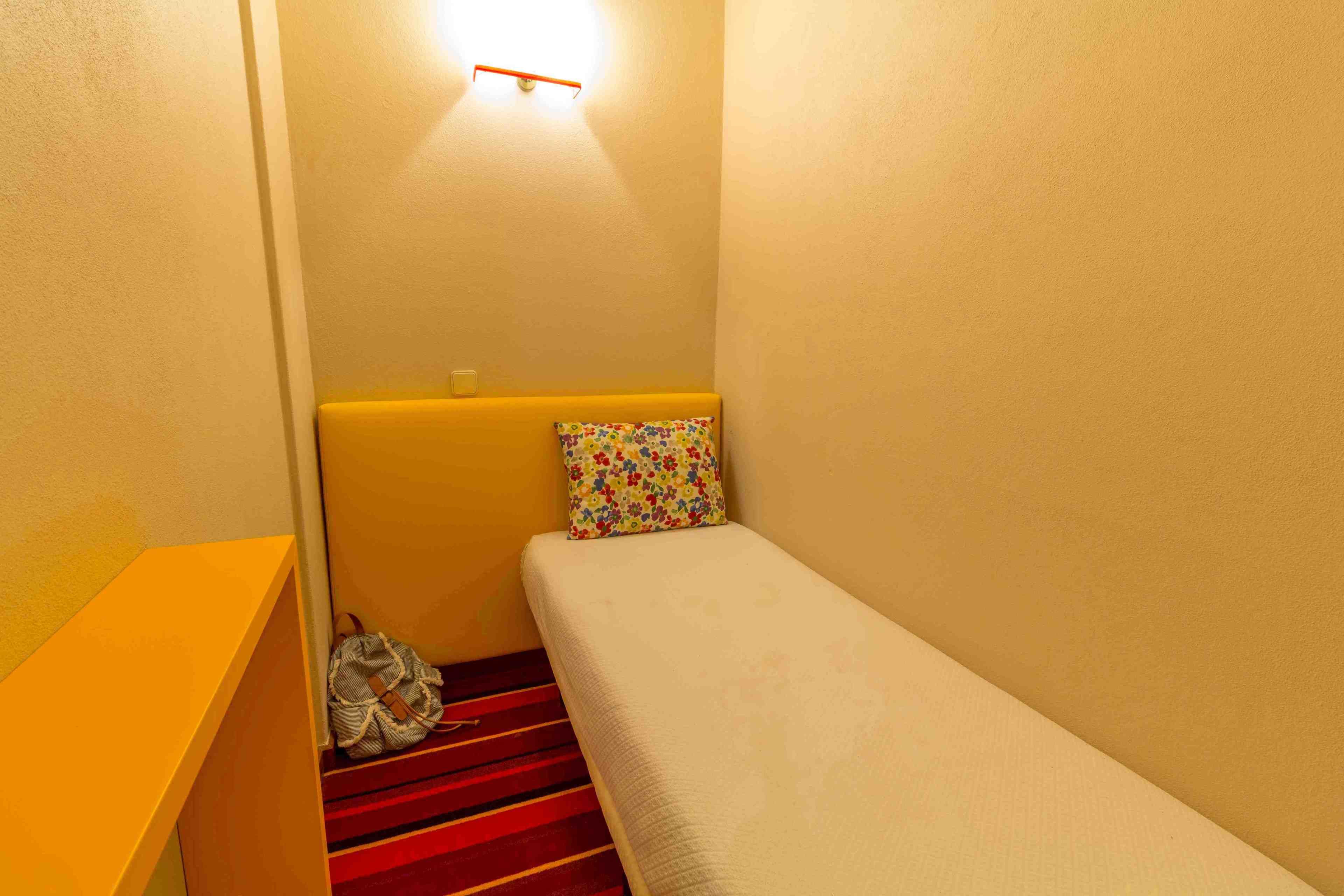 A small, single-bed room with a red-striped carpet, a yellow headboard, and a colorful pillow.