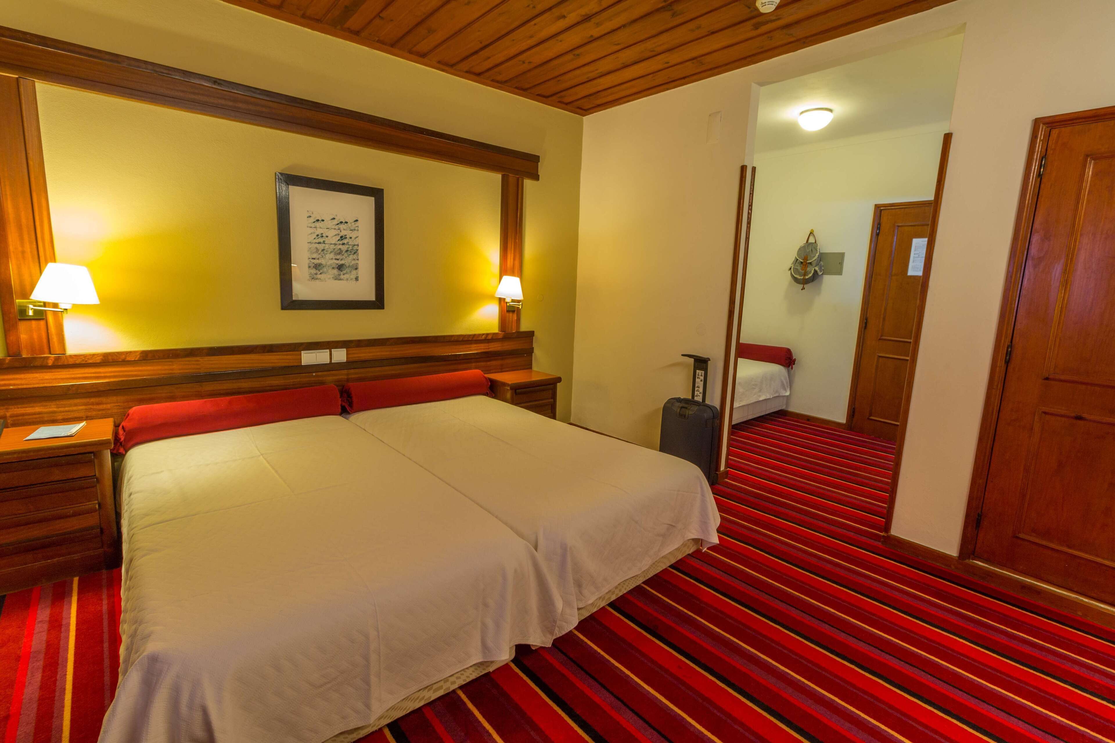 A hotel room with a large bed, wooden headboard, two bedside lamps, a red-striped carpet, and a suitcase.
