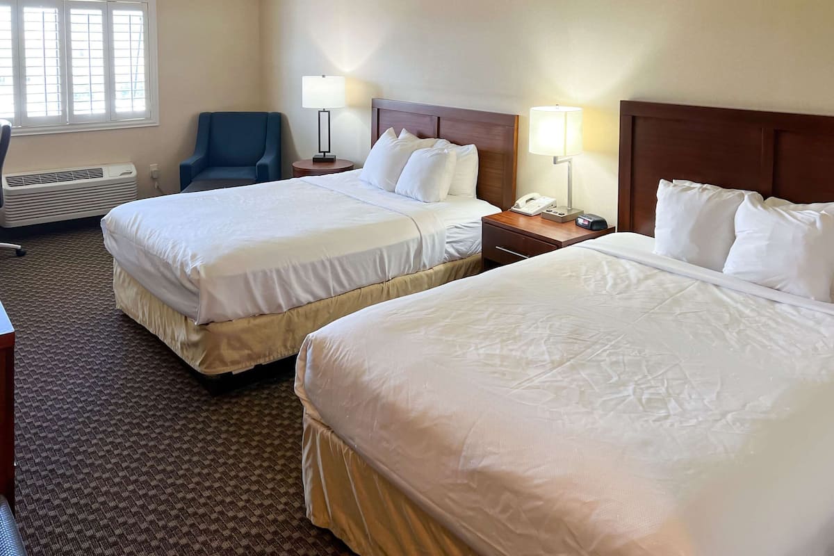 A hotel room with two beds, a desk, a chair, a lamp, and a bedside table.