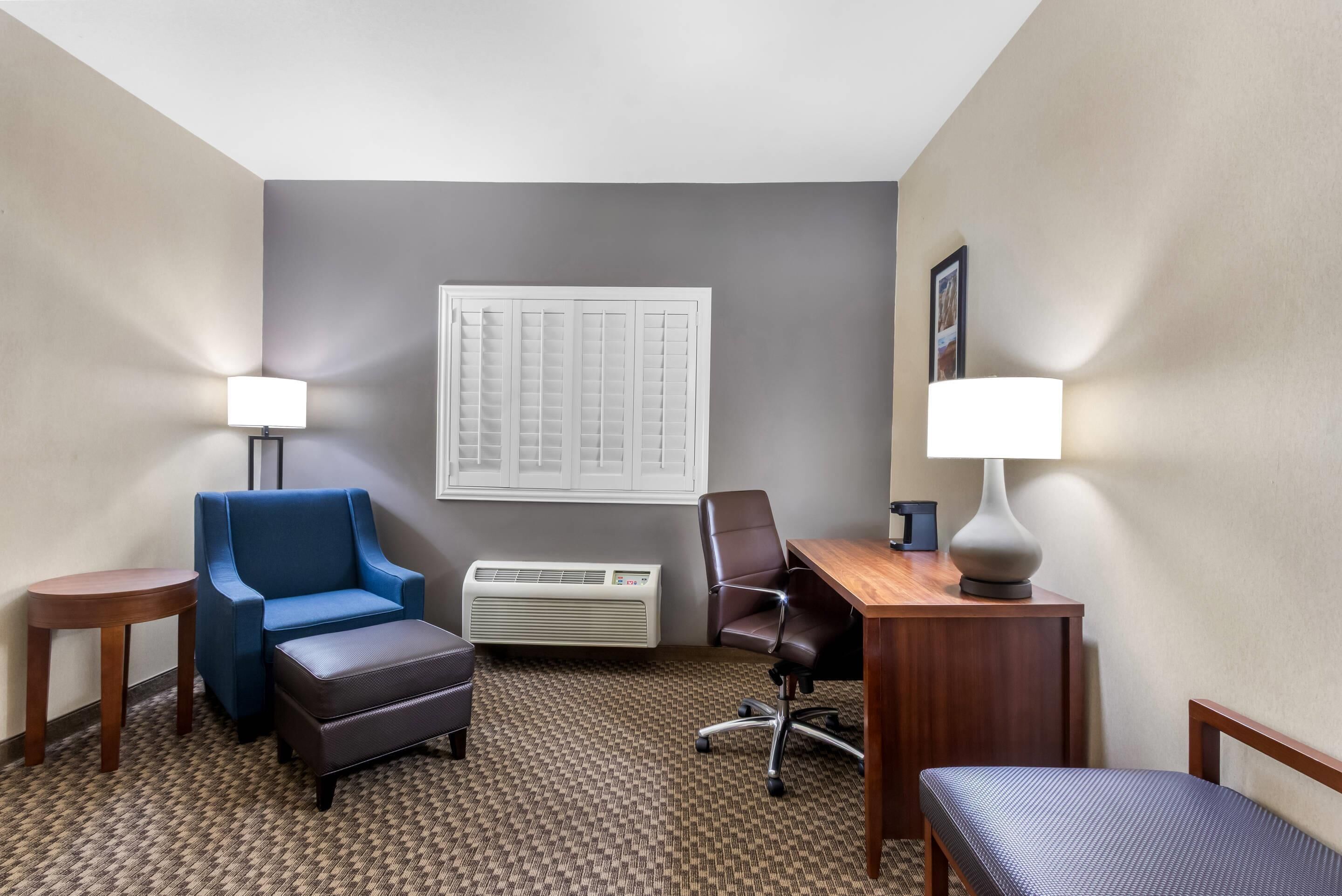 A hotel room with a blue armchair, a wooden desk, a brown leather ottoman, a black office chair, and a small wooden table.