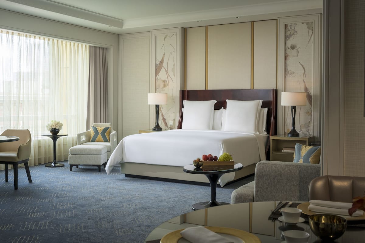 Une chambre d’hôtel avec un grand lit, deux tables de chevet avec des lampes, une table ronde sur laquelle se trouve un saladier rempli de fruits, une chaise et un canapé.