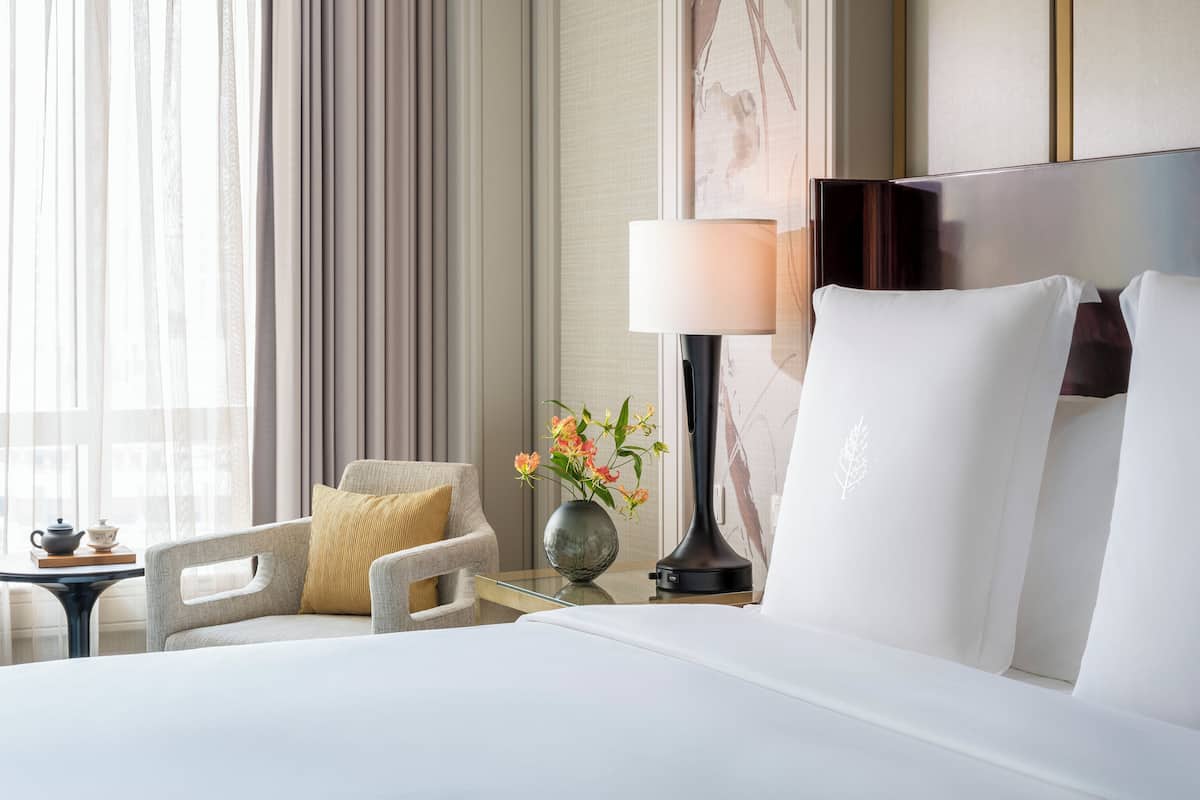 Une chambre d’hôtel avec un lit, une chaise, une table ornée d’un vase de fleurs et une lampe.
