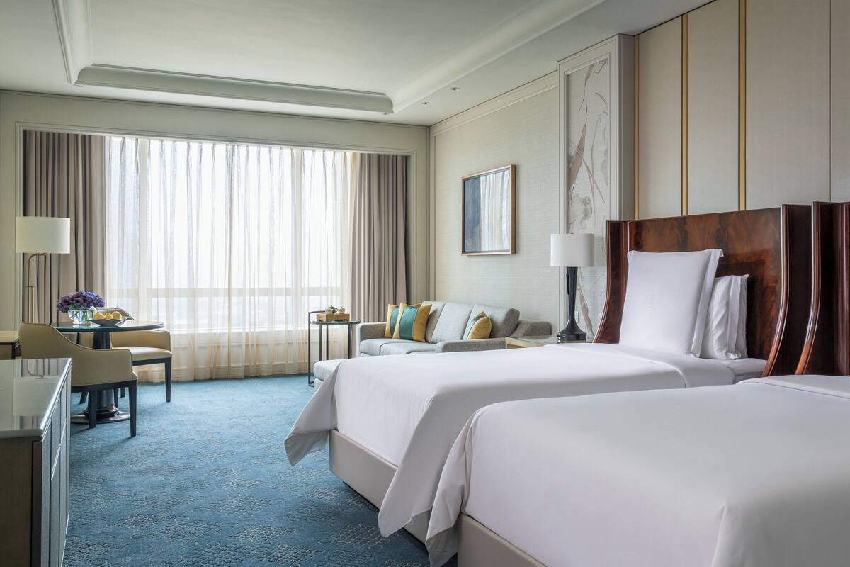 Une chambre d’hôtel avec deux lits, un bureau, une chaise et une vue sur l’extérieur grâce à de grandes fenêtres.