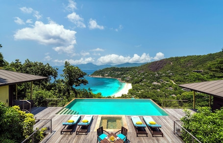 A pool with lounge chairs and a table, overlooking a beach and mountains.