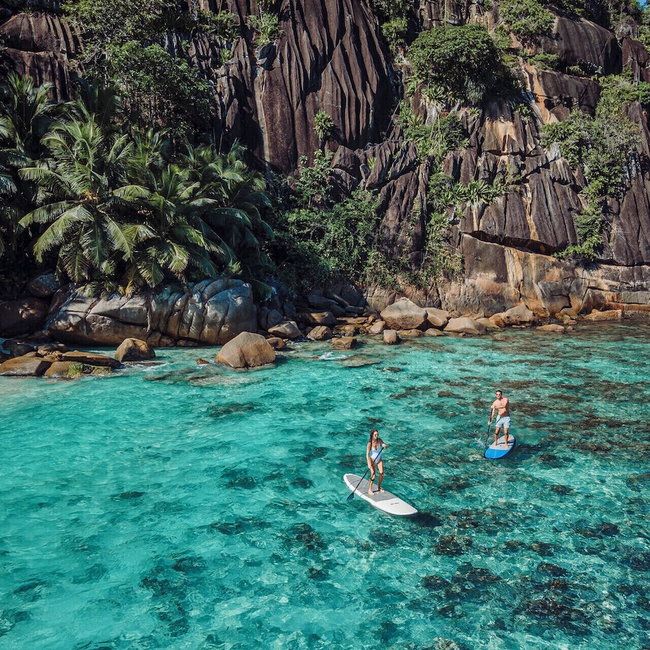 A tropical beach with clear turquoise water, palm trees, and rocky cliffs.