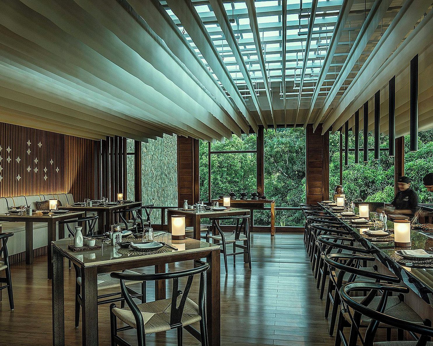A restaurant with a long dining area, wooden tables, and chairs, with a view of greenery outside.