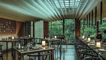 A restaurant with a long dining area, wooden tables, and chairs, with a view of greenery outside.