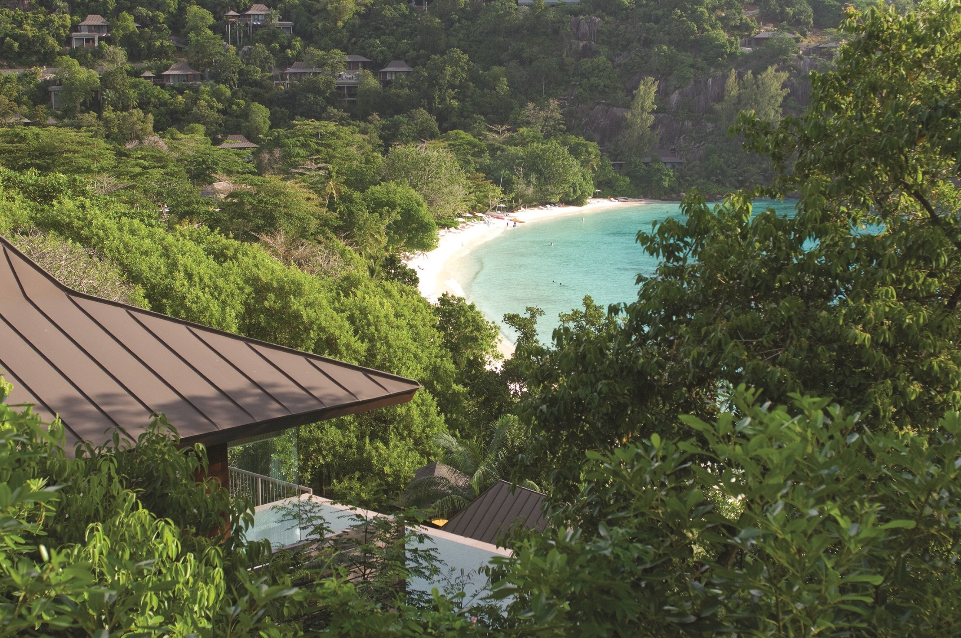 A coastal view with a beach, dense greenery, and a building with a sloped roof.