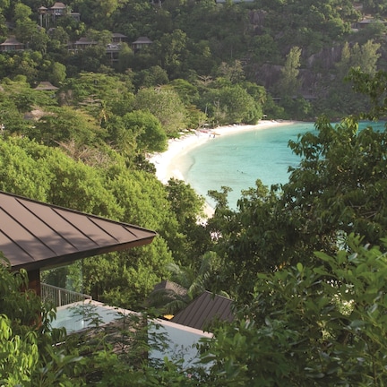 A coastal view with a beach, dense greenery, and a building with a sloped roof.
