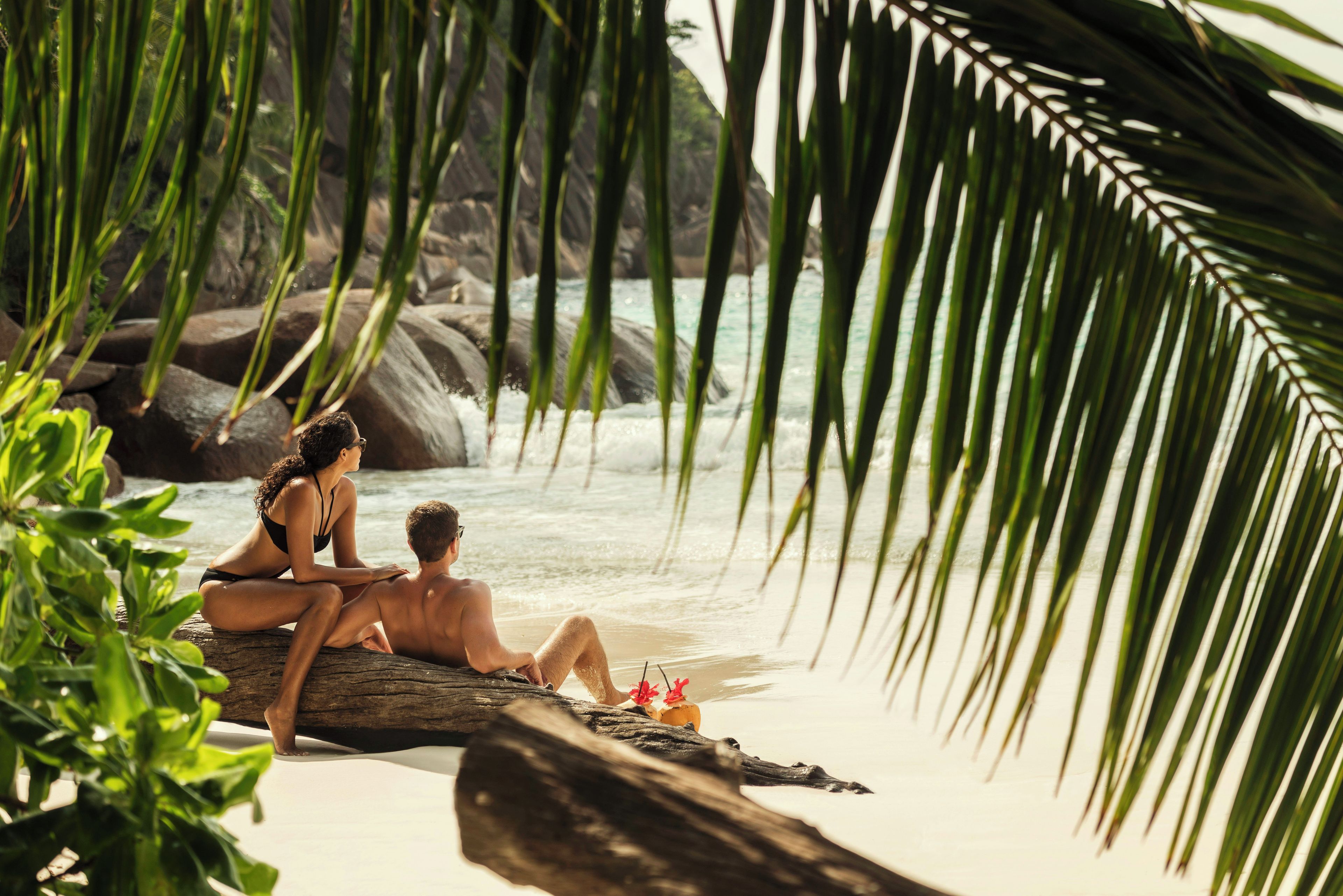 A couple sitting on a rock by the water, surrounded by greenery.