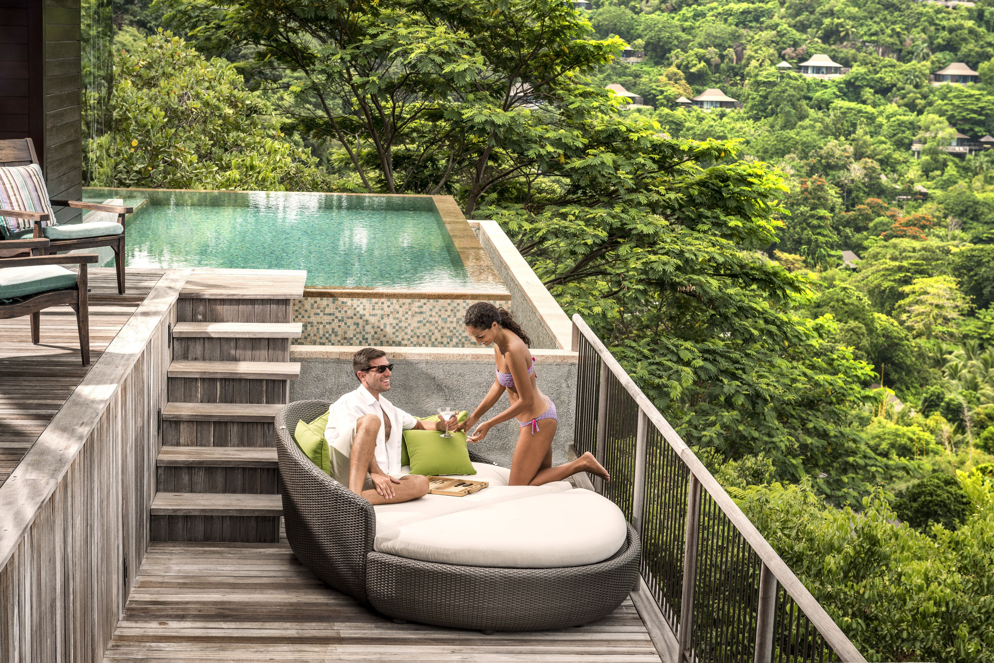A wooden deck with a pool, a wicker lounge chair, and a view of a lush forest.