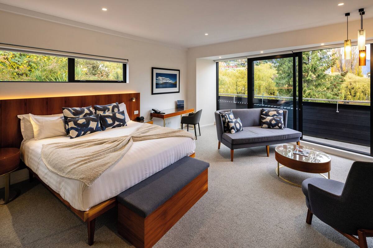 A modern bedroom with a large bed, a sofa, a desk, and a balcony with a view of greenery.