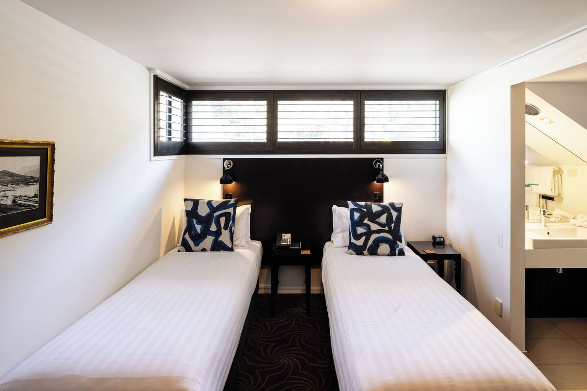 A hotel room with two single beds, a small bedside table with a phone, a window with blinds, and a bathroom visible through an open door.