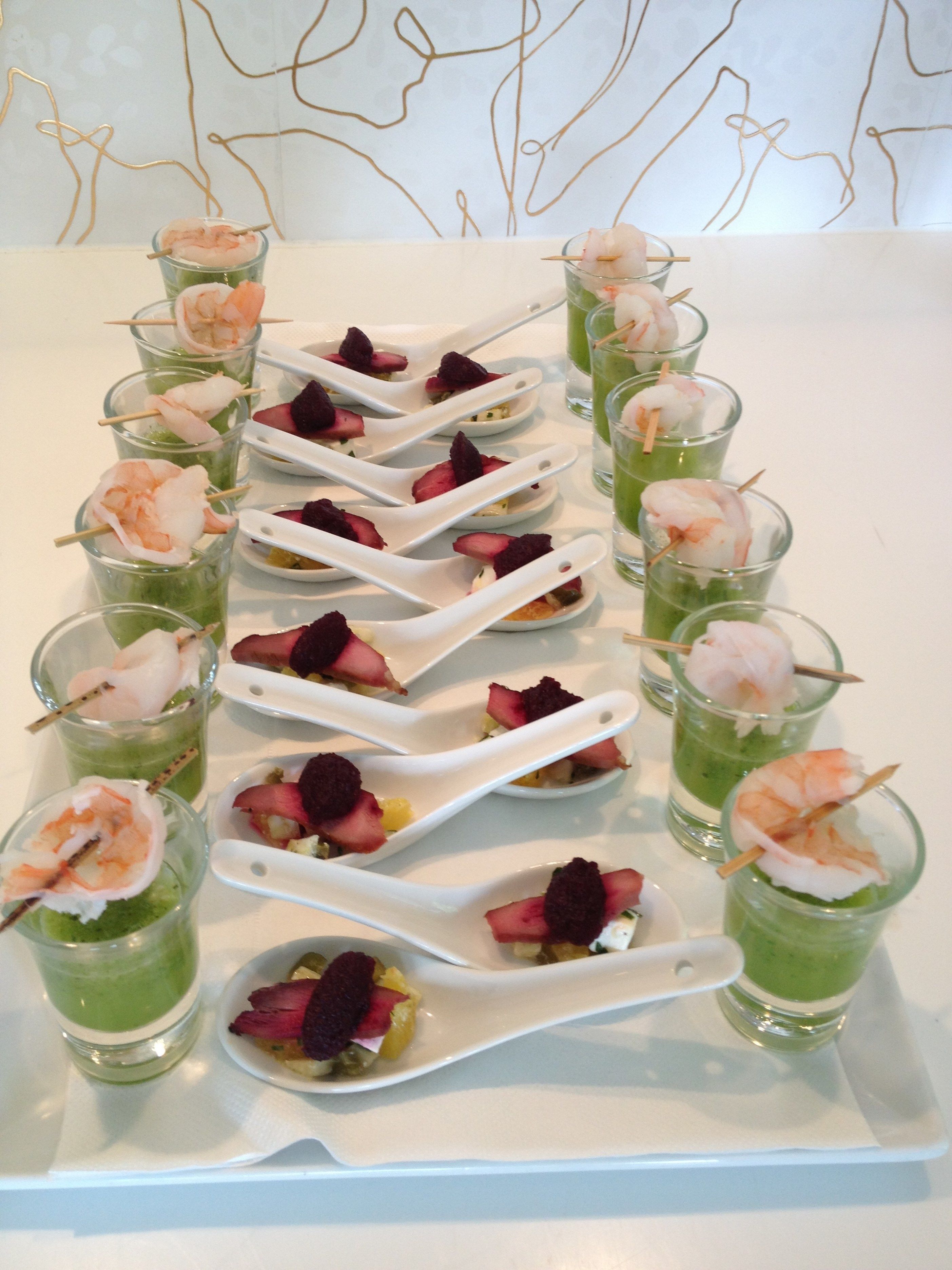 A table with shot glasses containing green beverages, garnished with shrimp and berries, and white spoons.