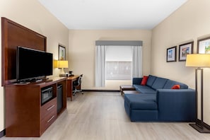 A hotel room with a flat-screen TV, a desk with a lamp, a blue sofa, and a window with curtains.
