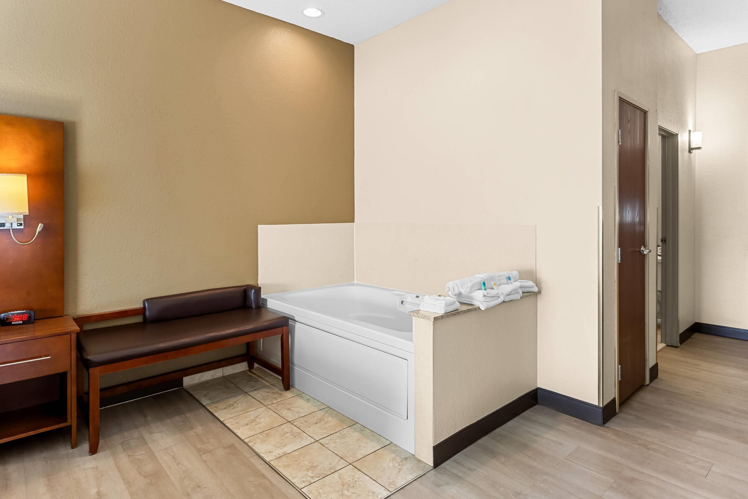Una habitación de hotel con cama, un banco, una mesita y la puerta del baño.