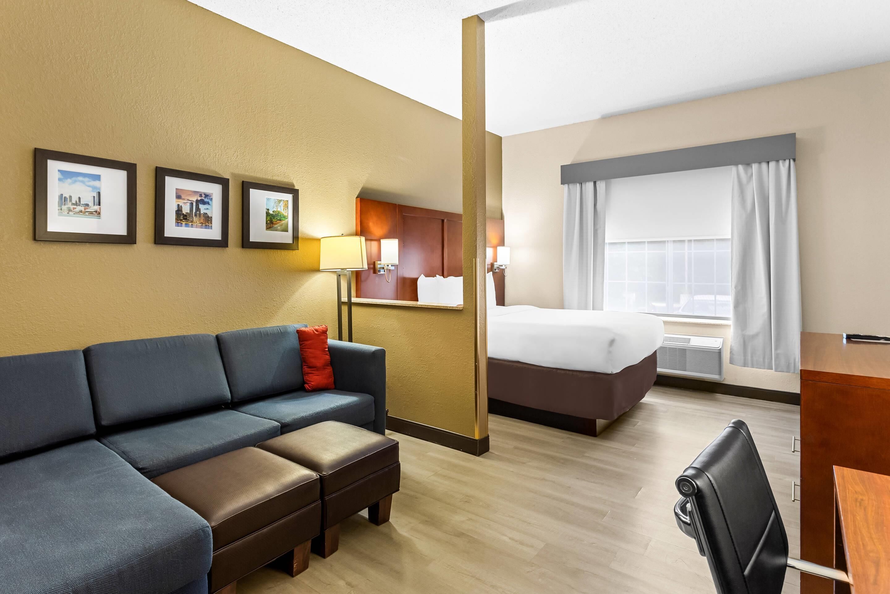 Ett hotellrum med en soffa, en säng, ett skrivbord och en stol. På väggen hänger inramade tavlor och det finns ett fönster med gardiner.